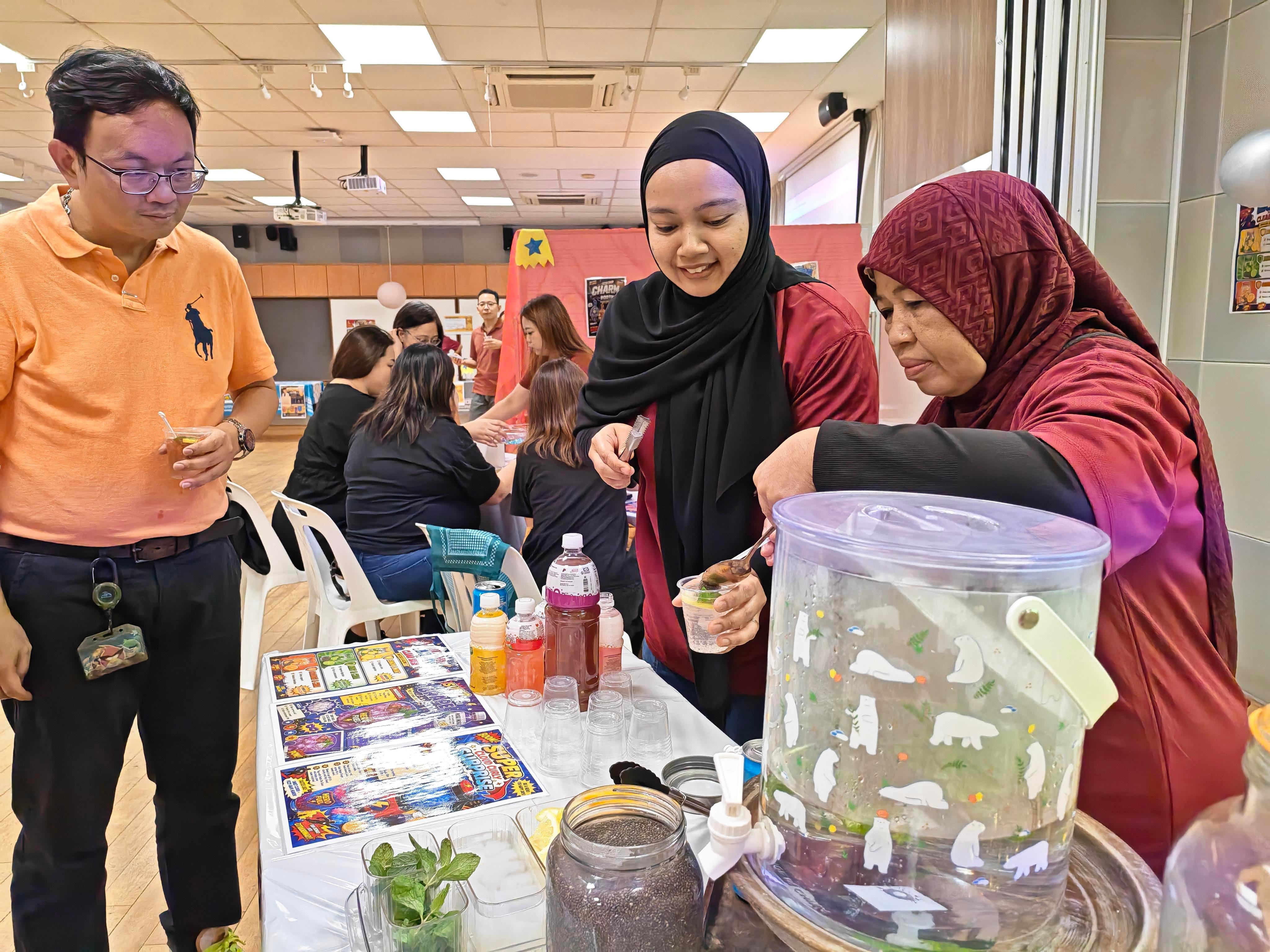 Two parent volunteers prepare drinks at a booth set up with colourful beverage bottles, cups, and fresh ingredients. A staff member looks on as the volunteers assemble the drinks, with other teachers enjoying activities at booths in the background.
